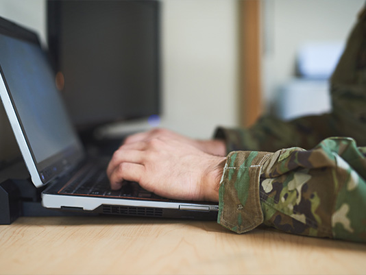 A person in military fatigues typing on a laptop